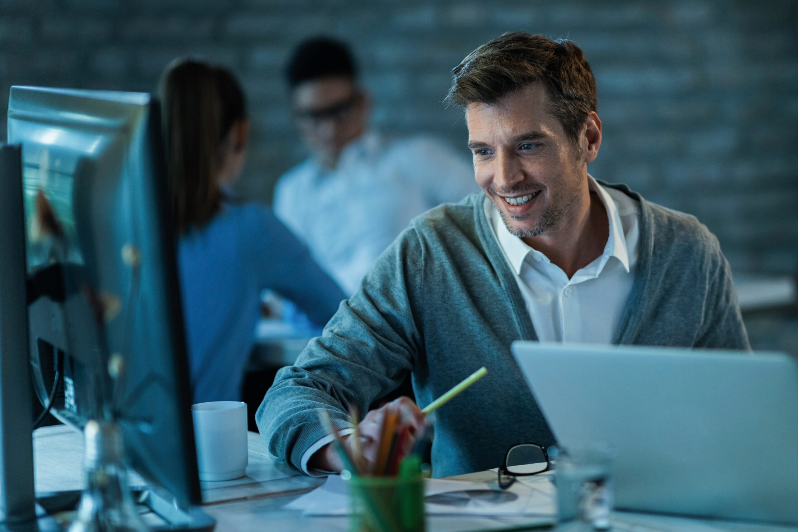 Happy male entrepreneur using desktop PC while working in the office. There are people in the background.